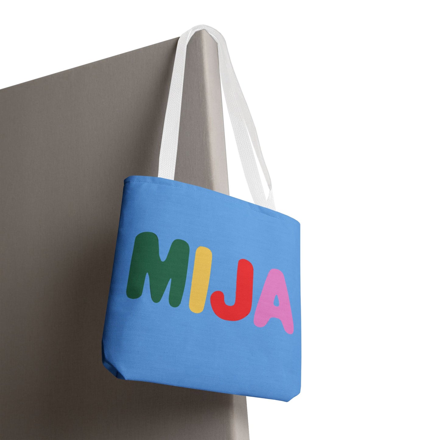 Blue tote bag with colorful letters spelling 'MIJA' hanging on a white surface.
