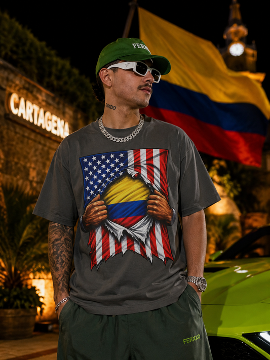 Man wearing a t-shirt with a design of an American flag and Colombian flag, standing in front of a Colombian flag and 'Cartagena' sign.