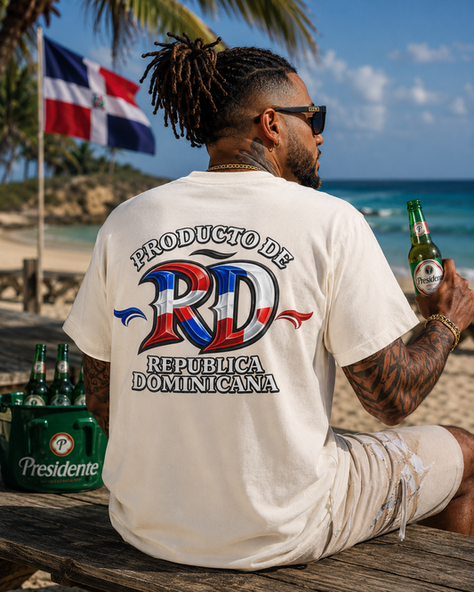 A man sitting on a beach bench seen from behind, wearing a cream-colored t-shirt with a large graphic on the back featuring the text 'PRODUCTO DE REPUBLICA DOMINICANA' and a stylized 'RD' in the Dominican flag colors. He is holding a green beer bottle, with other bottles and a cooler on the bench, and the Dominican Republic flag flying in the background.