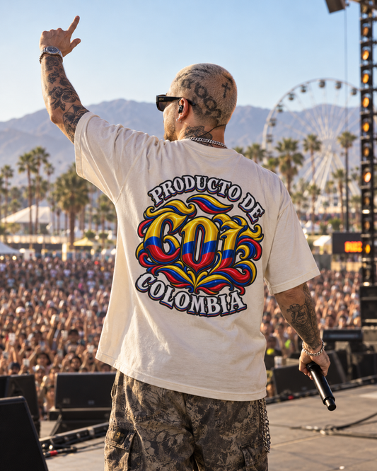 A man stands on a concert stage with his back to the camera, wearing a white graphic T-shirt that reads 'PRODUCTO DE COLOMBIA' with a large colorful Colombian flag-themed design on the back. The background shows a large crowd, palm trees, mountains, and a Ferris wheel under a clear blue sky.