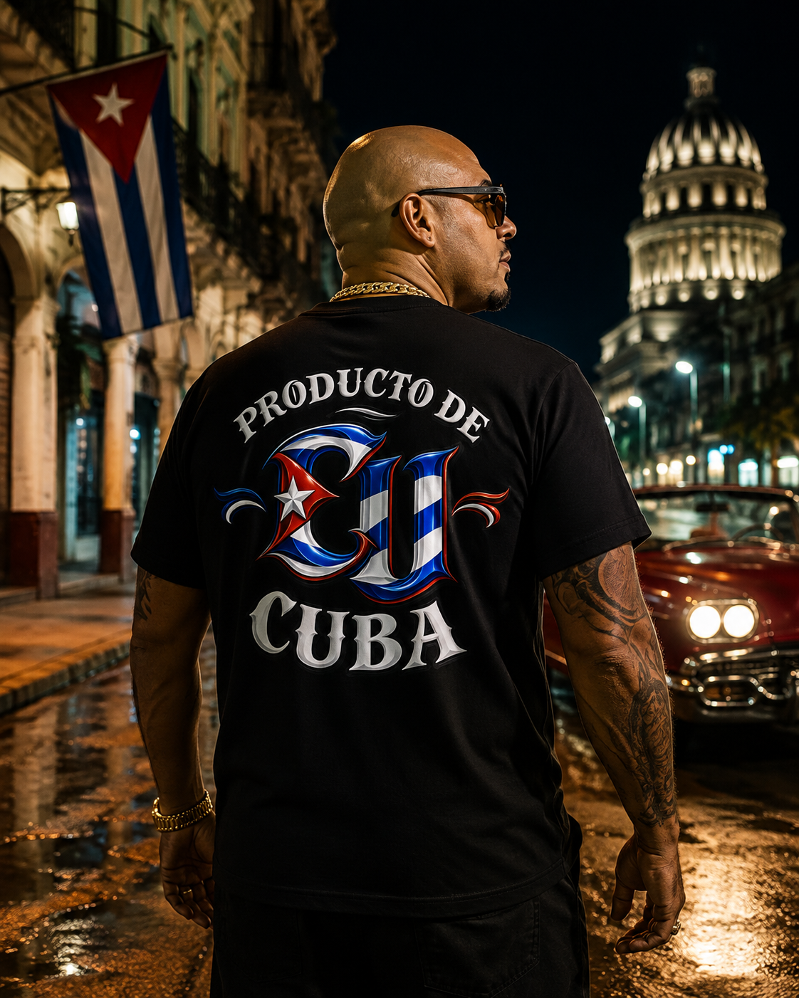 A man wearing a black t-shirt with a large Cuban-themed graphic on the back, standing on a wet street at night with a Cuban flag and a domed capitol building in the background.