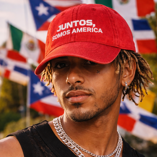 Man wearing a red cap with 'Juntos Somos America' text, standing in front of various flags.