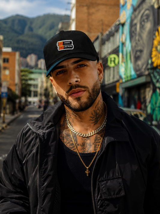Man wearing a black cap and jacket with tattoos, standing in an urban street setting.