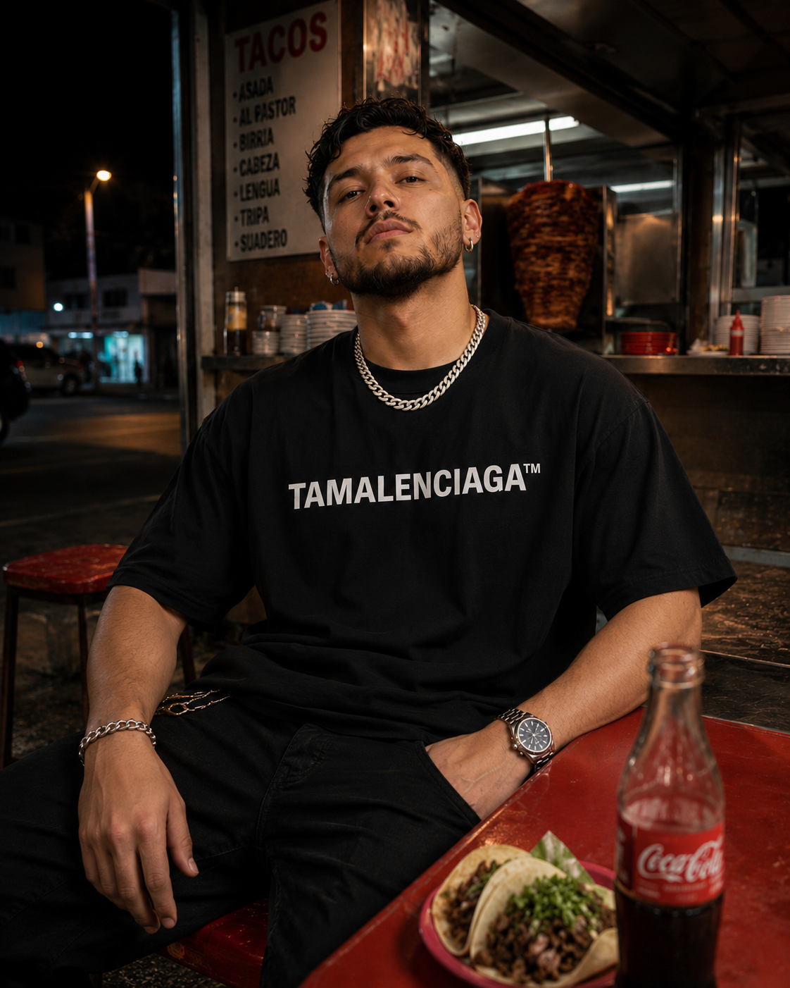 Man wearing a black 'TAMALENCIAGA' t-shirt sitting at a table with food and a Coca-Cola bottle in a casual setting.