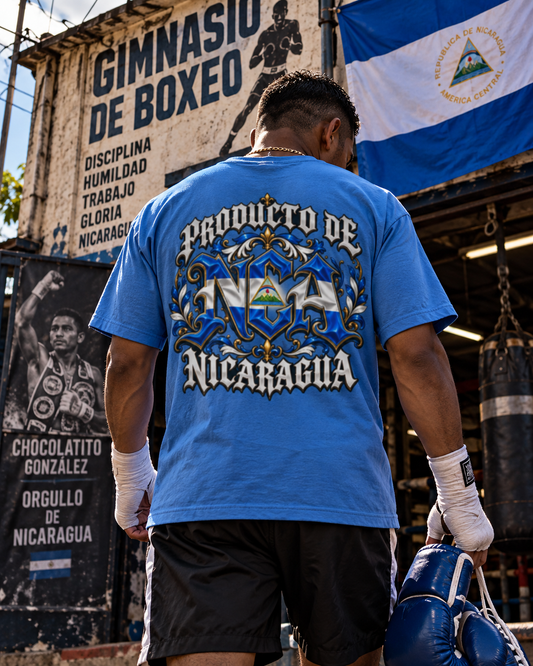 A man viewed from behind wearing a bright blue t-shirt with a large graphic on the back that reads 'Producto de Nicaragua' in white and blue. He is standing in a boxing gym holding blue boxing gloves, with a Nicaraguan flag and a boxing poster in the background.