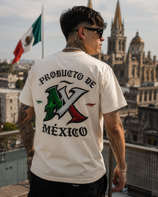 A man wearing a cream-colored t-shirt with a large graphic on the back that reads 'PRODUCTO DE MÉXICO' with a stylized 'MX' in the colors of the Mexican flag, standing on a rooftop with a cityscape and Mexican flag in the background.