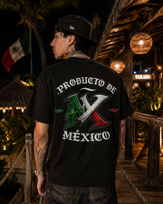 A man wearing a black t-shirt with a large 'PRODUCTO DE MÉXICO' graphic featuring the Mexican flag colors on the back, photographed in a tropical night setting with palm trees and warm lights.