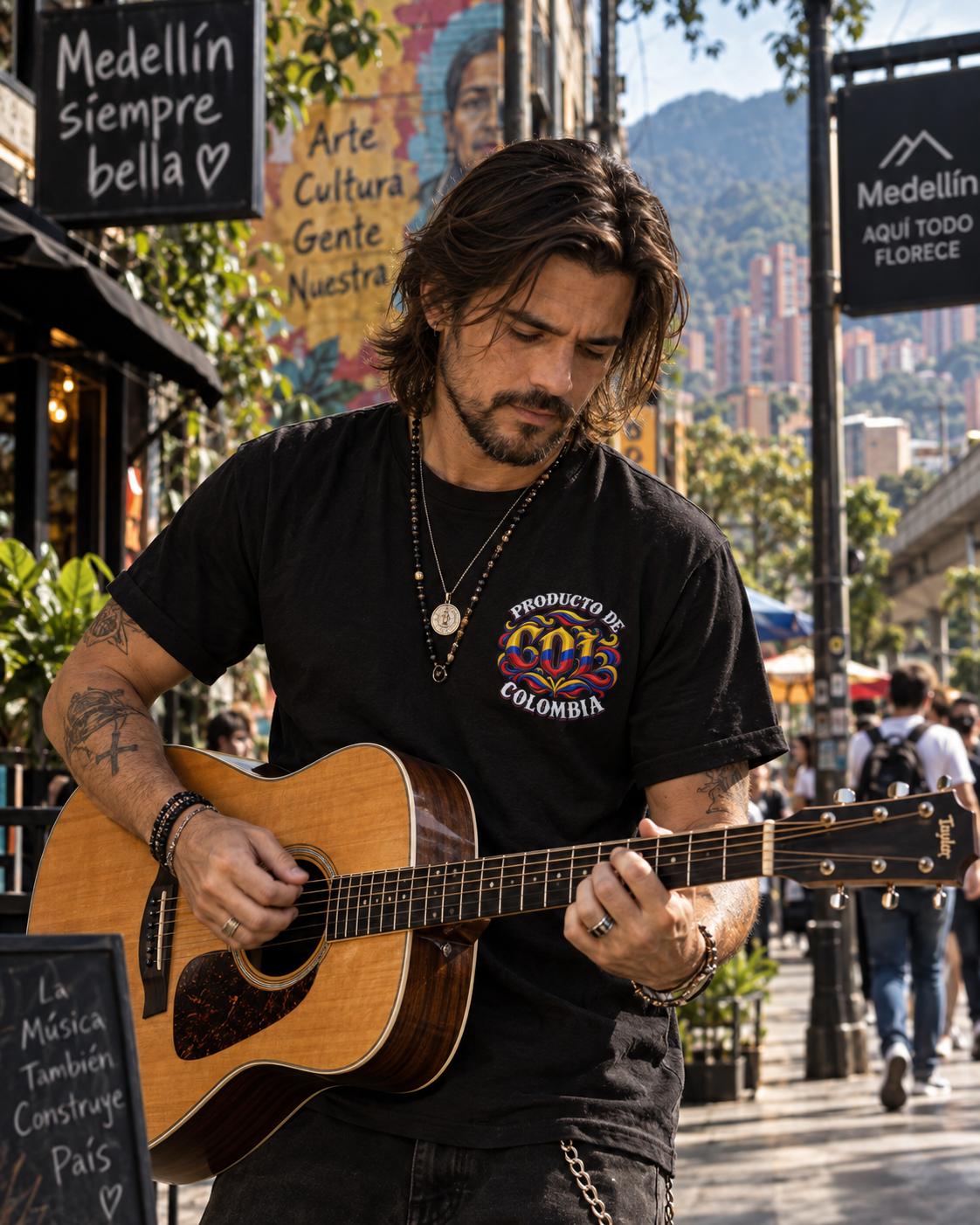A man with long dark hair and a beard playing an acoustic guitar on a city street. He is wearing a black graphic t-shirt with a colorful circular logo that reads 'PRODUCTO DE COLOMBIA' and 'MEDIELLIN'.