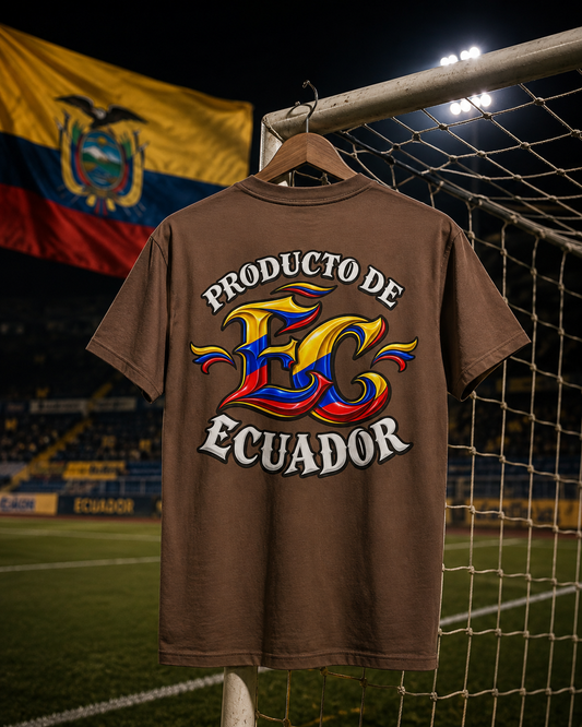 A brown short-sleeve t-shirt hanging on a wooden hanger in front of a soccer goal net at night, featuring a large graphic on the back with the text 'PRODUCTO DE ECUADOR' and a stylized 'EC' logo in yellow, blue, and red, with the Ecuador flag blurred in the background.